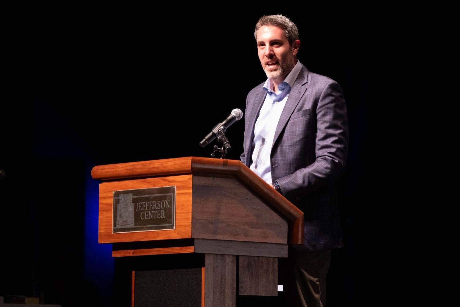Professional man in suit speaks at wooden podium on dark stage at formal event.