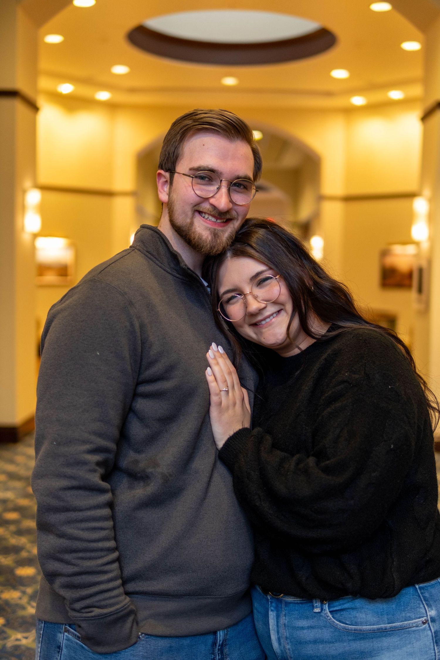 Happy engaged couple embracing and smiling in warmly lit hotel lobby, woman showing diamond engagement ring.