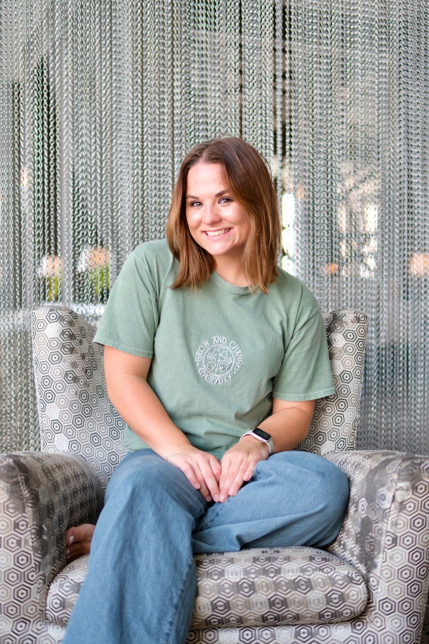 Smiling woman in green t-shirt sitting in patterned armchair against shimmering backdrop.
