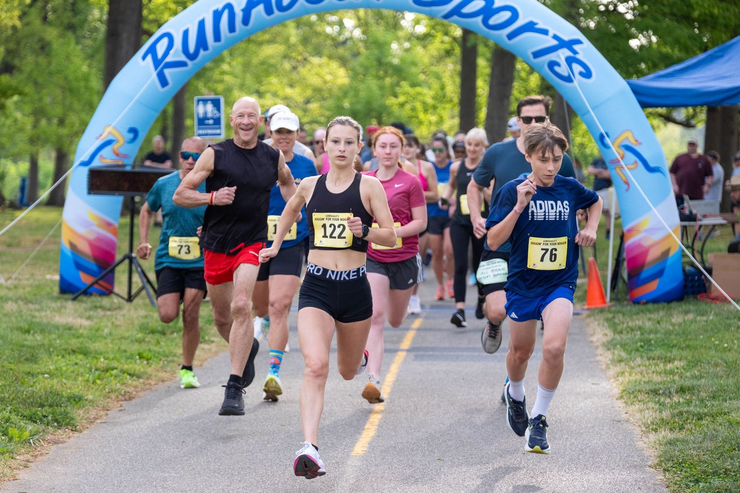 Runners race under a RunAbout Sports inflatable arch at an outdoor 5K road race event in a park.