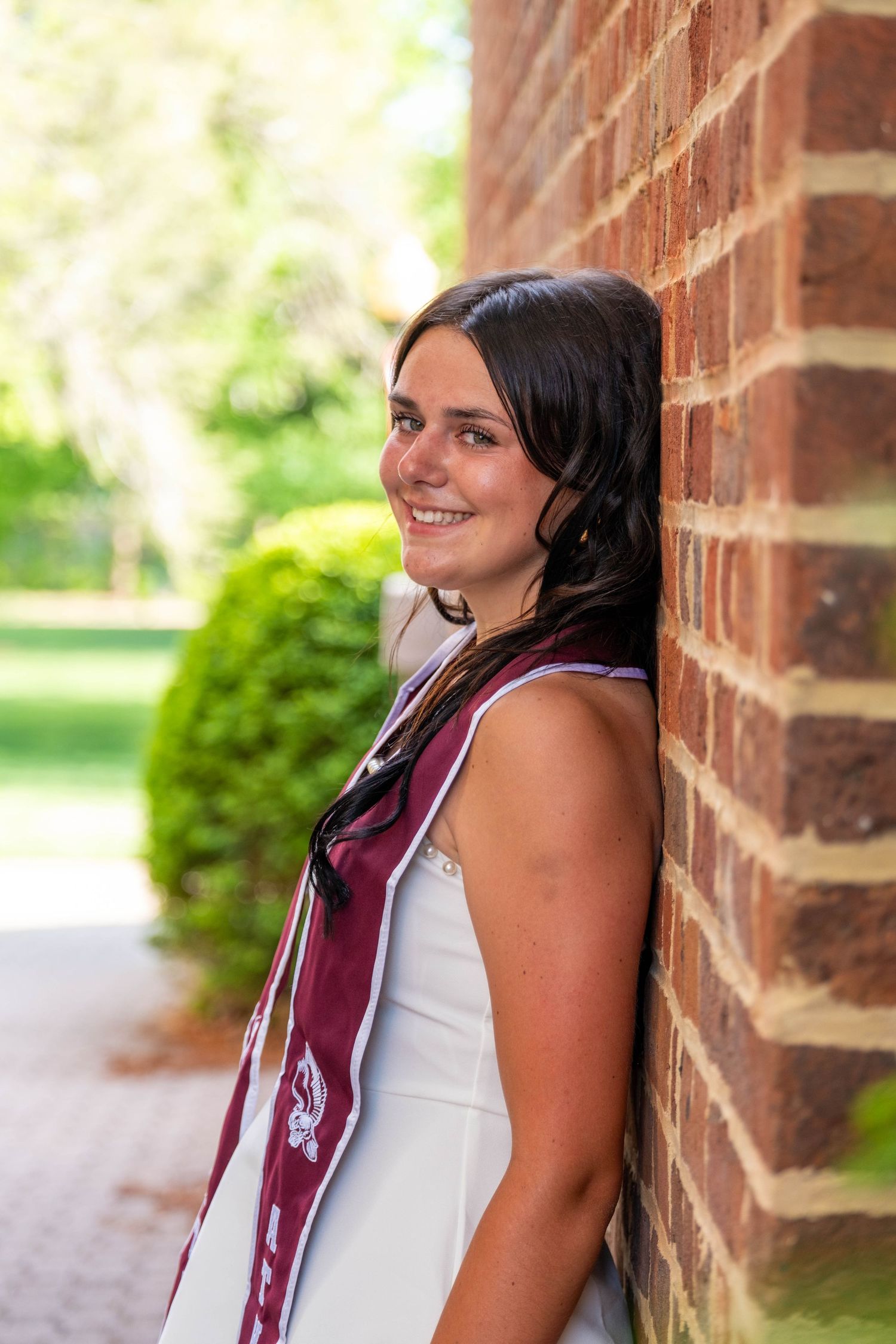 Smiling young woman in white dress with maroon graduation stole leaning against brick wall outdoors.