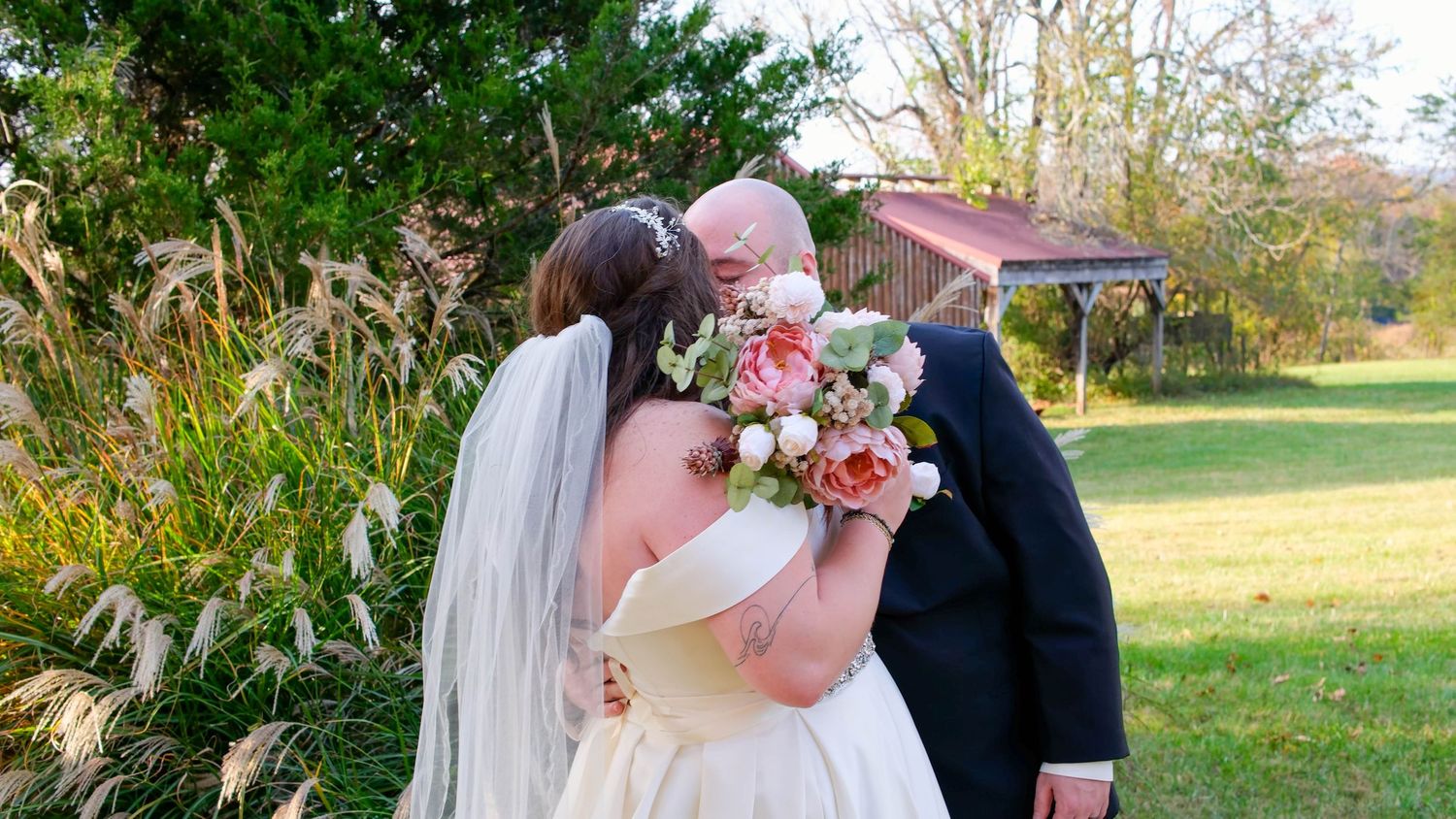 Bride and groom sharing a kiss outdoors with bride holding pink floral bouquet wearing white gown and veil.