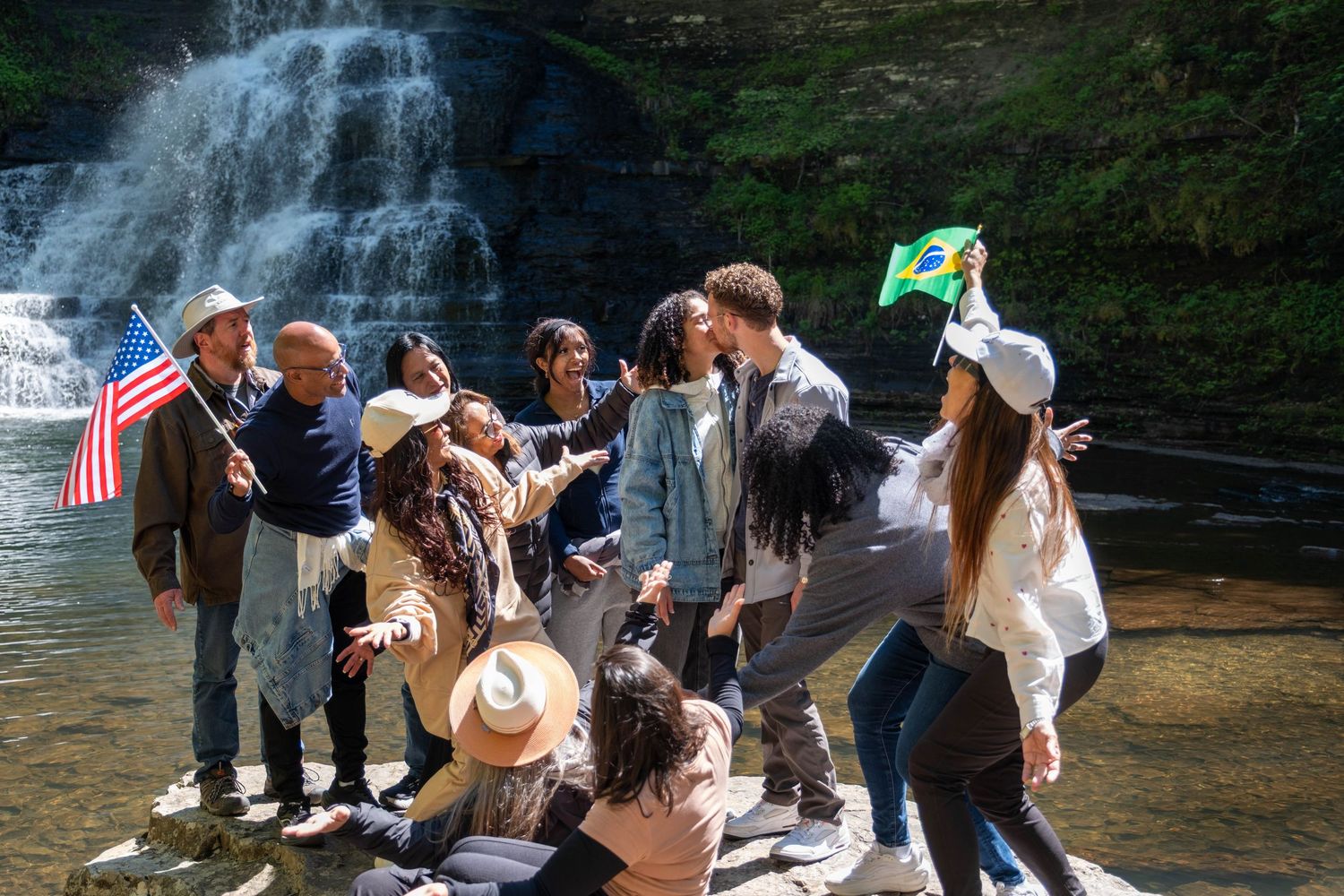 Diverse group of friends celebrating with American and Brazilian flags near a scenic waterfall on a sunny day.