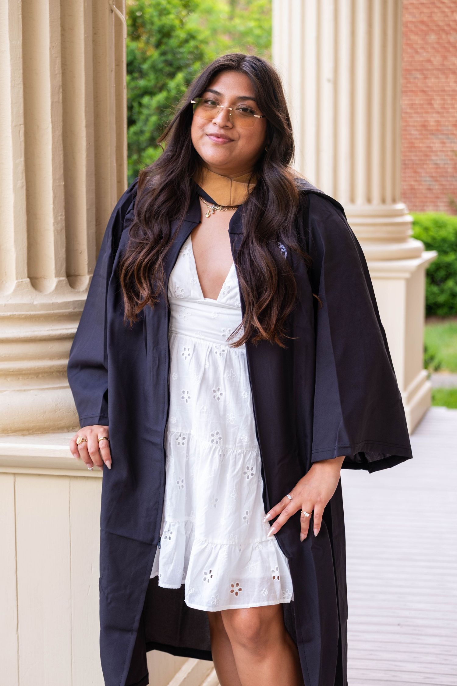 Young woman in graduation gown over white dress poses by columns outdoors, smiling confidently.