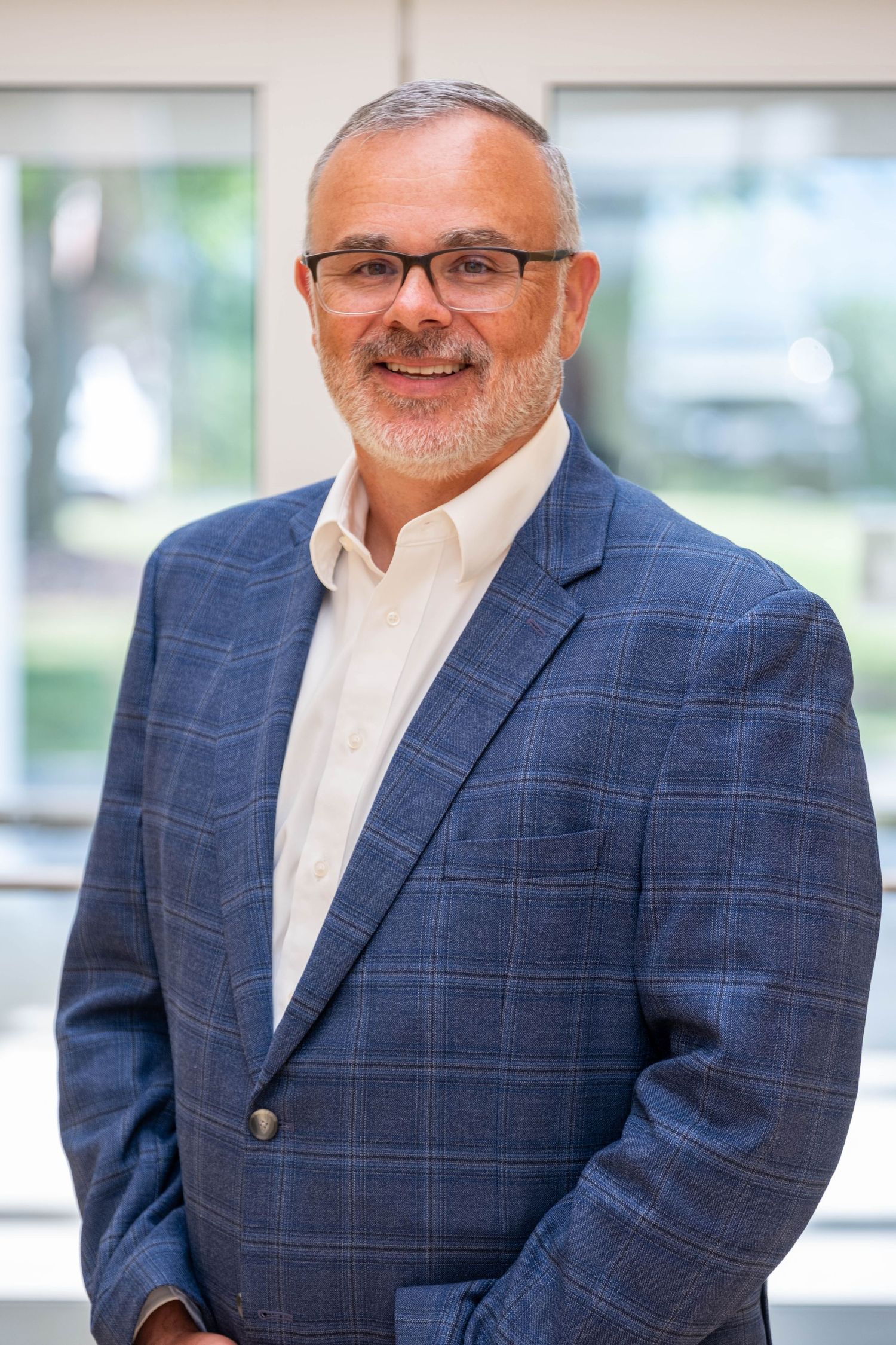 Professional man in blue plaid blazer and glasses smiling confidently in a bright modern office setting.