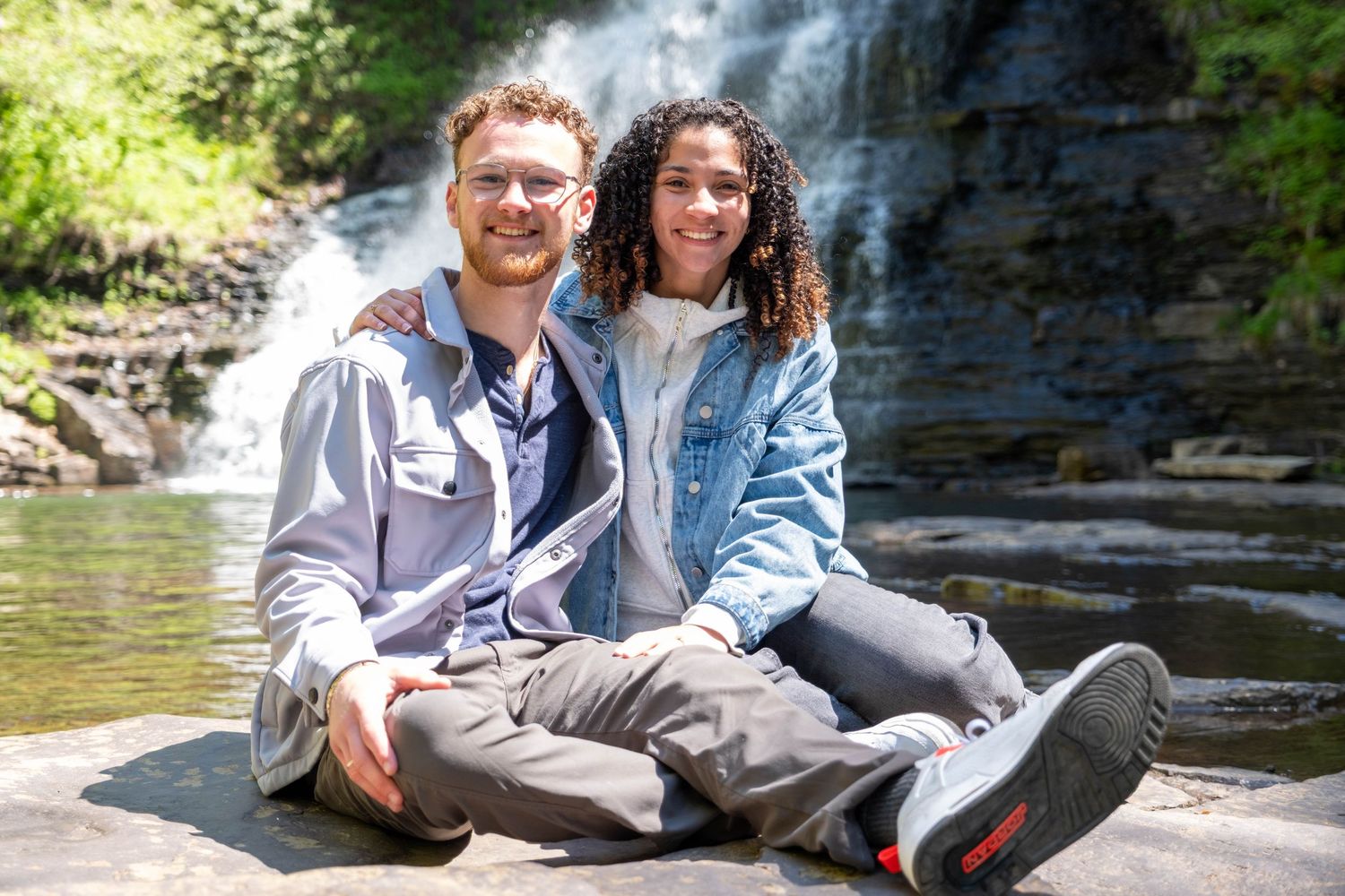 Couple smiling together on rocks in front of a scenic waterfall surrounded by lush greenery on a sunny day.