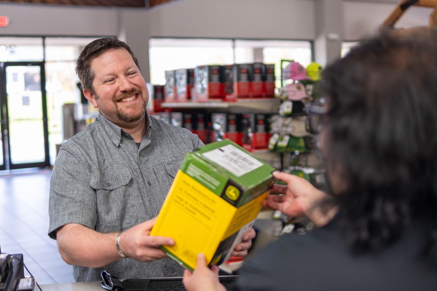 A smiling store employee hands a John Deere product box to a customer at a retail counter.