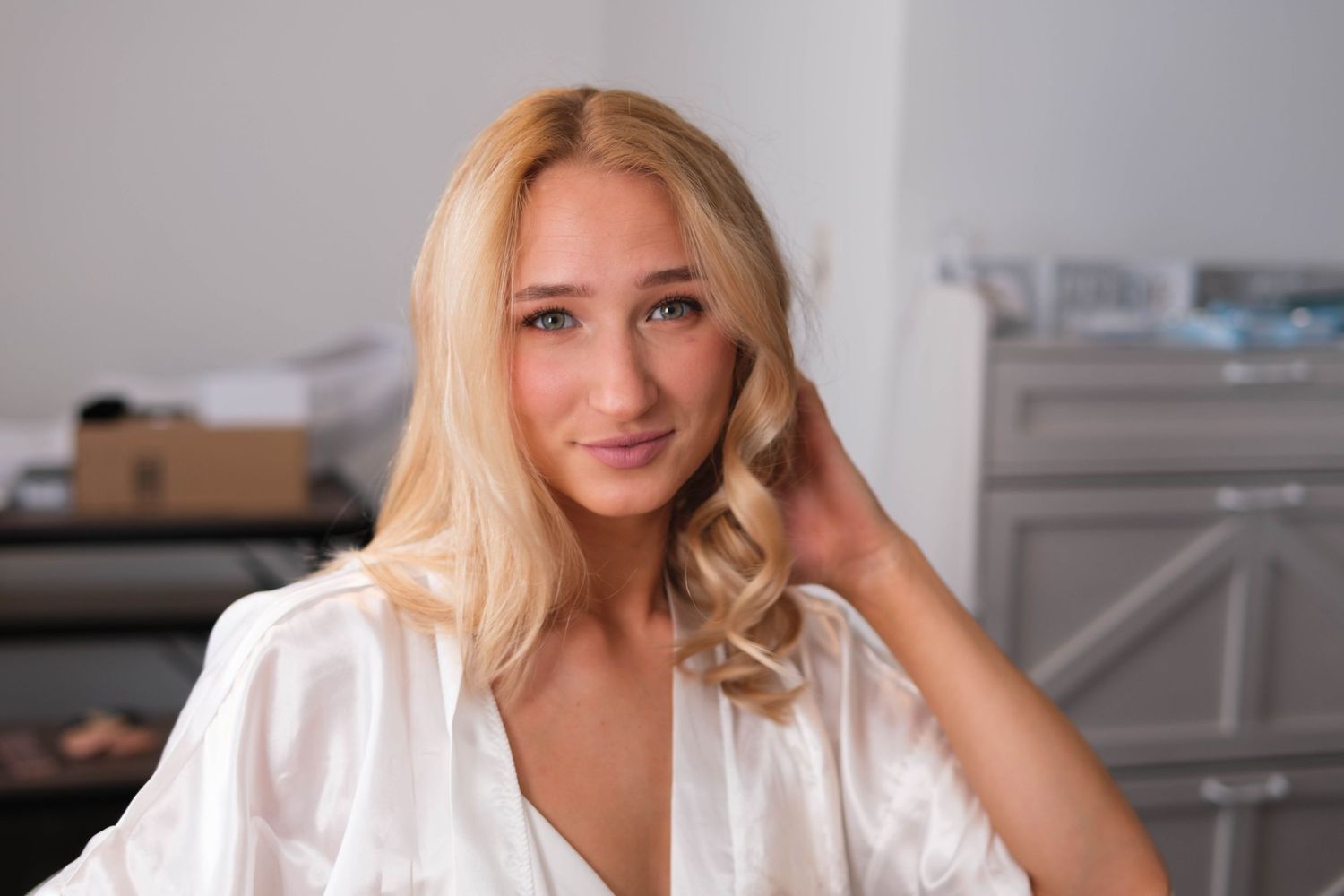 Beautiful young blonde woman in white satin robe smiling softly in a bright modern room.