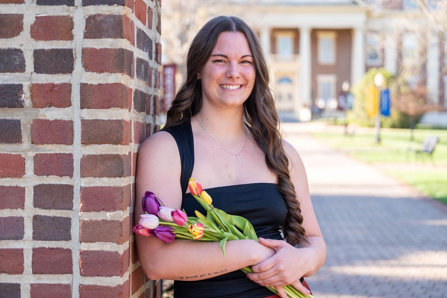 Smiling young woman holding colorful tulip bouquet while leaning against brick wall on campus.