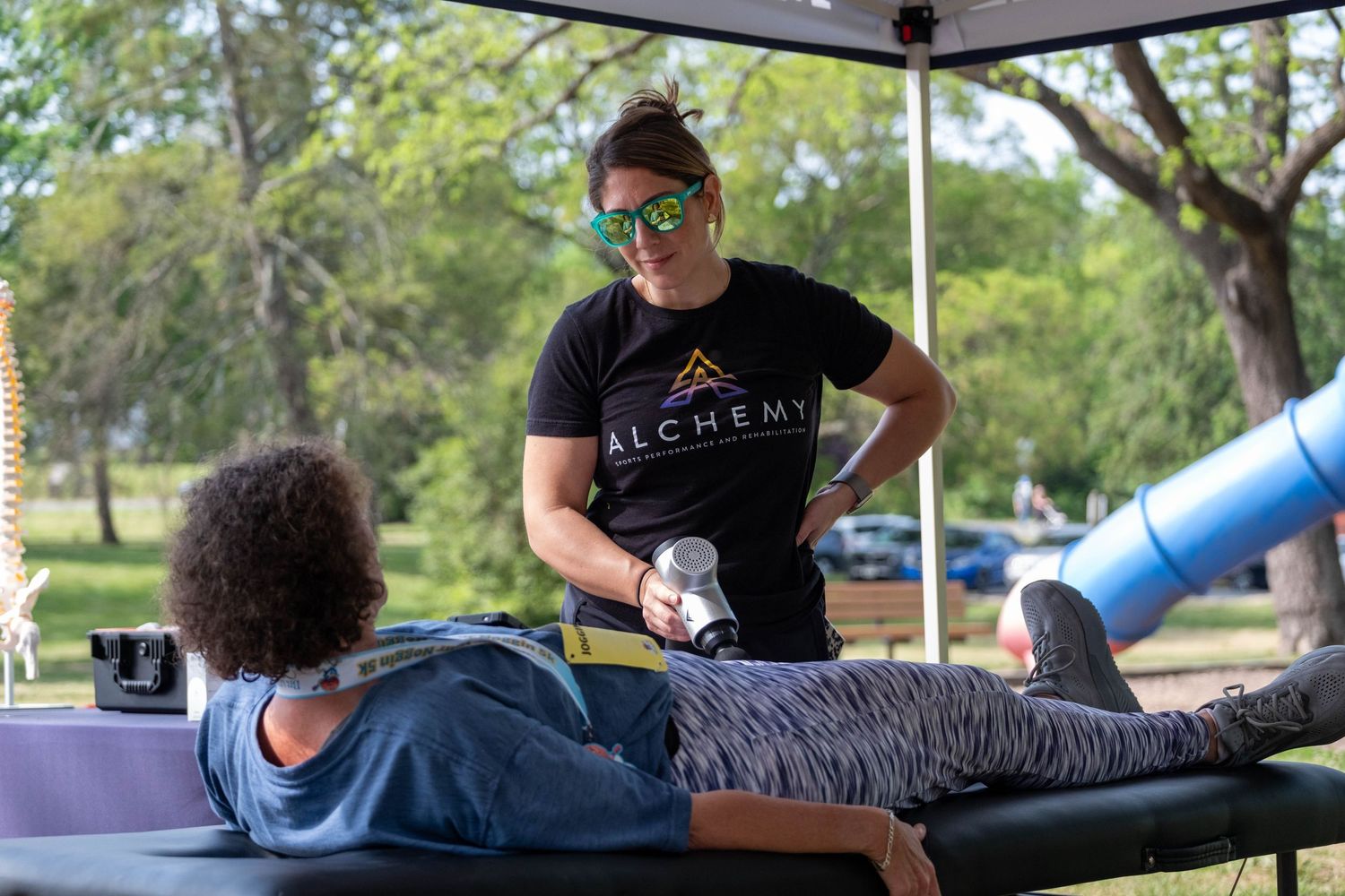 Alchemy therapist using massage tool on client lying on outdoor table at park event.