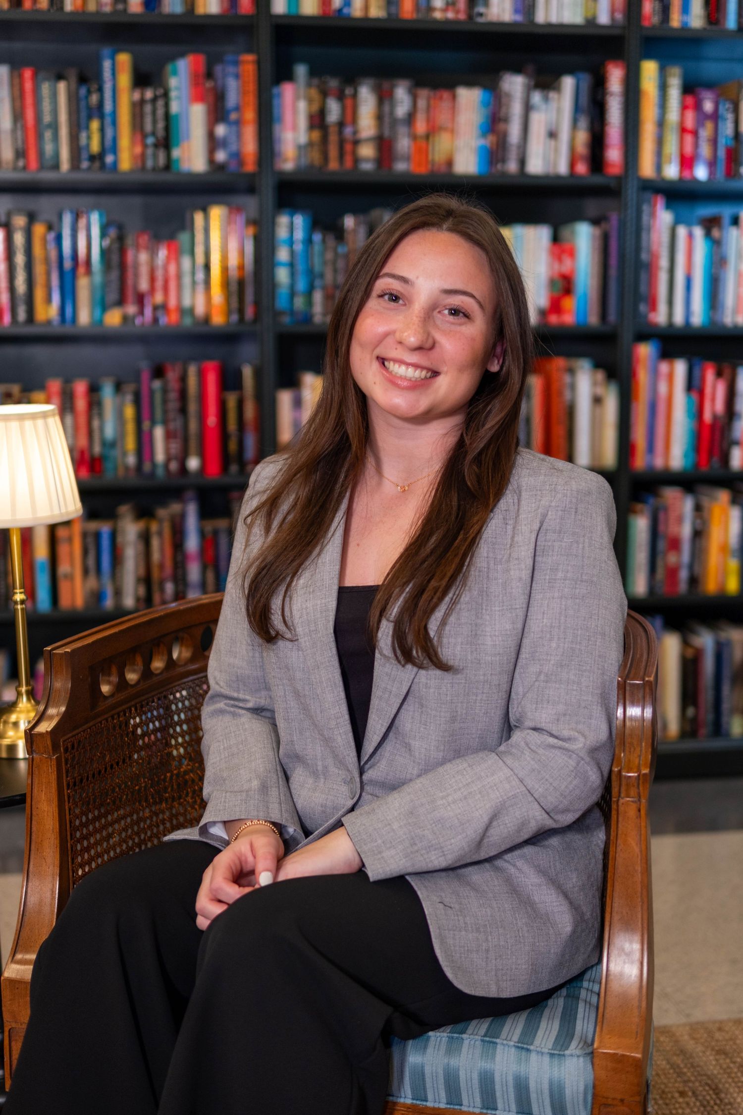 Young woman in gray blazer sitting in wooden chair smiling in front of colorful bookshelf library background.