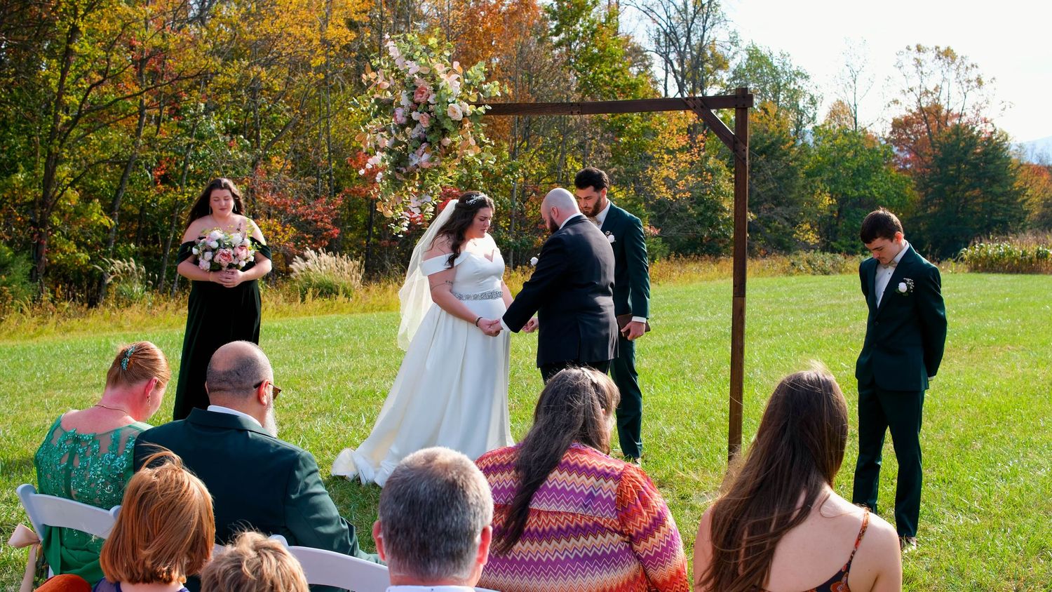 A bride and groom exchange vows under a wooden arch in an outdoor fall wedding ceremony with seated guests.
