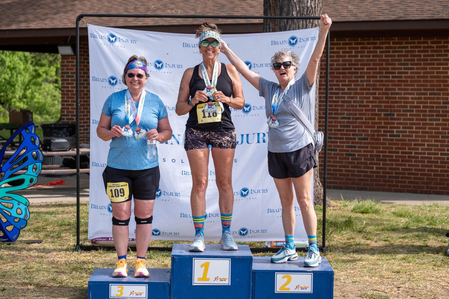 Three women stand on a race podium holding medals at an outdoor running event on a sunny day.