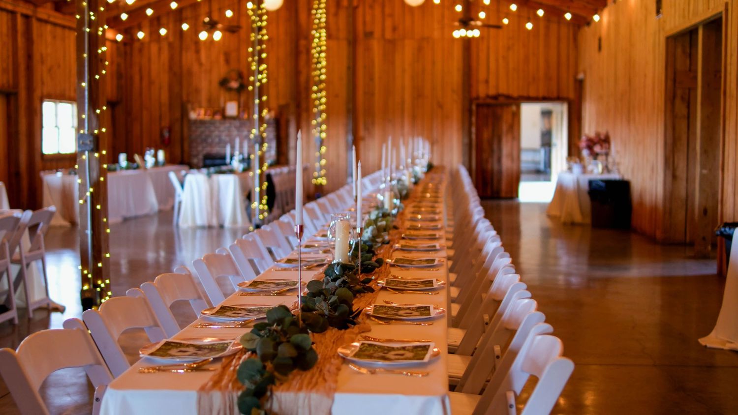 An elegant barn wedding reception hall decorated with string lights and a long table adorned with greenery centerpieces.