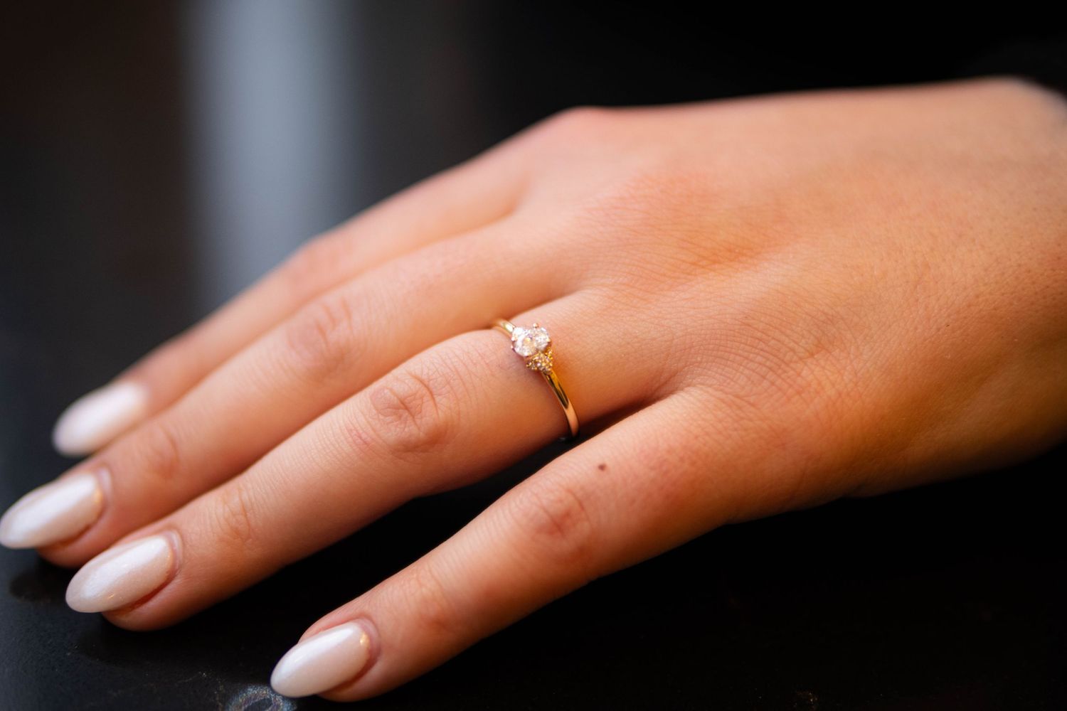 Close-up of elegant gold diamond engagement ring on hand with white manicured nails on dark background.