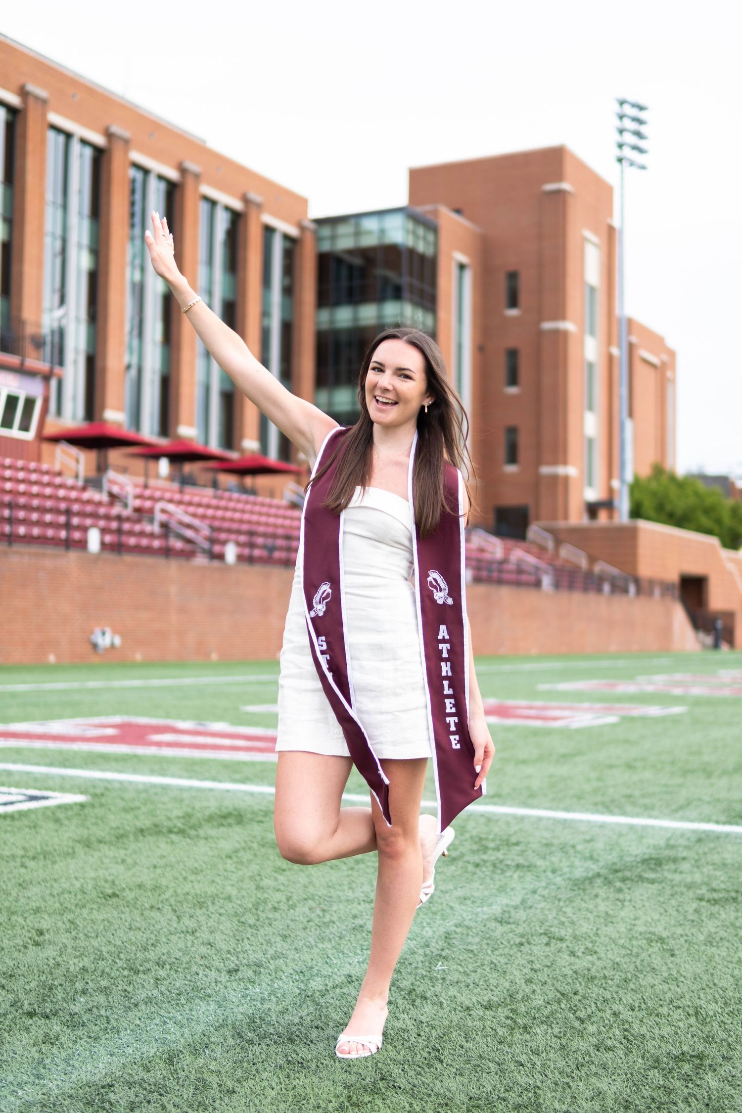 Female graduate in white dress and maroon athlete sash posing on college football field.