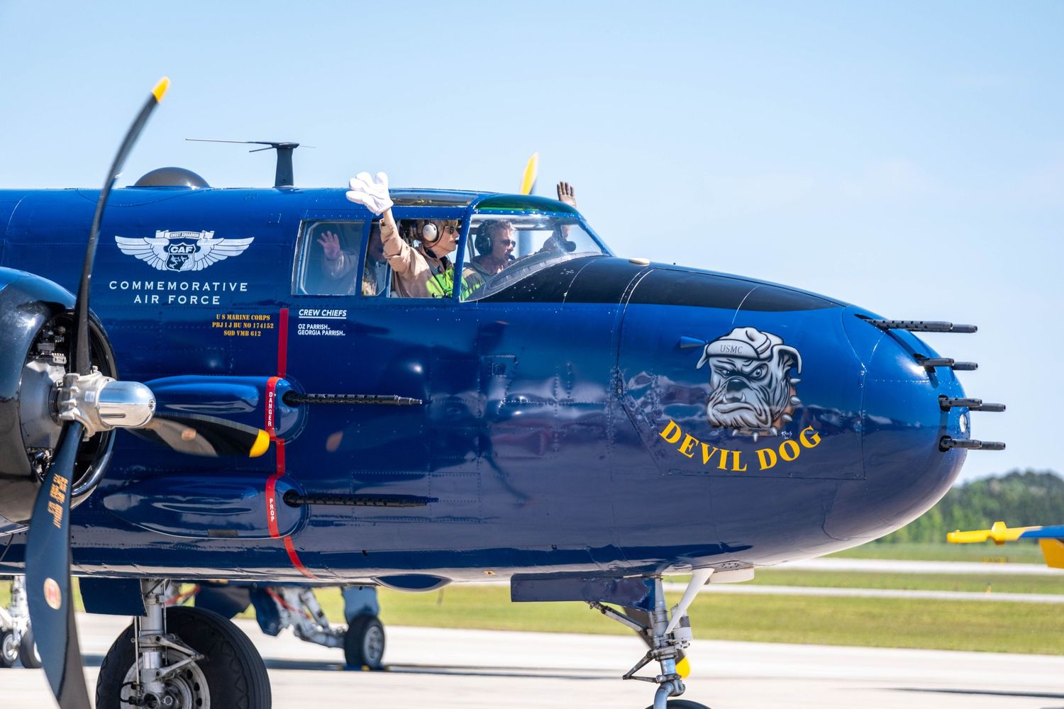 Blue Commemorative Air Force Devil Dog aircraft with pilot waving on a sunny runway.
