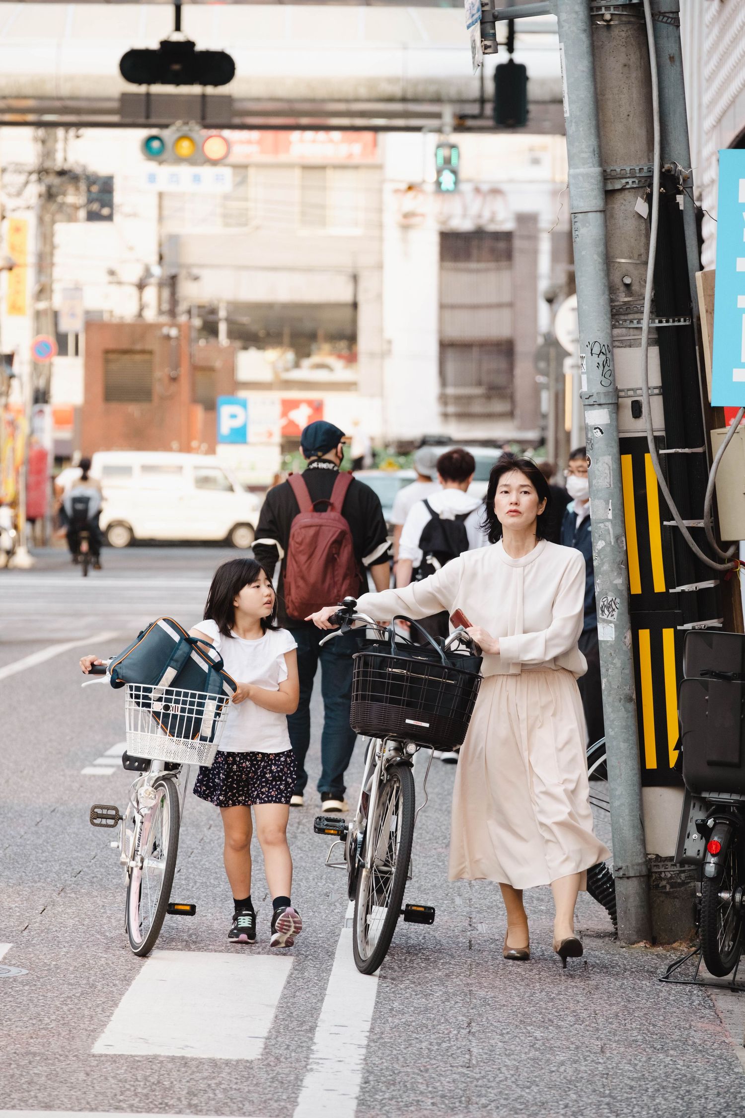 A woman and young girl wait at a crosswalk with bicycles on a busy urban street in Japan.