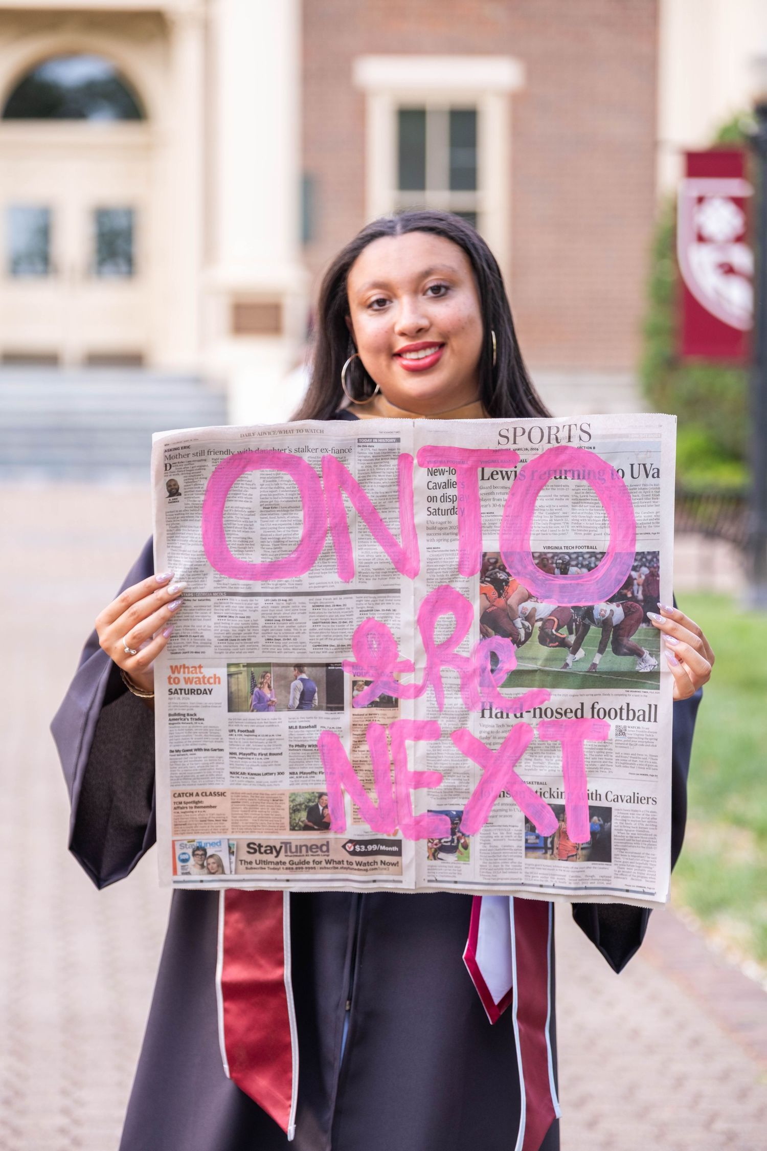 Graduate in cap and gown holds newspaper painted with 'Onto the Next' celebrating college graduation outdoors.