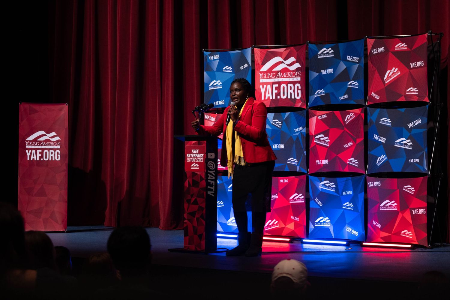 Speaker standing at YAF.org conference podium wearing red blazer and yellow scarf on stage.