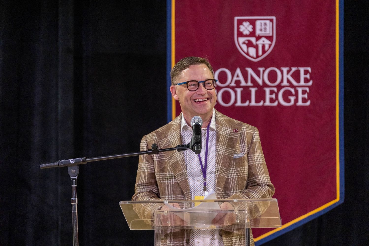 Smiling man in plaid blazer speaking at Roanoke College podium event.