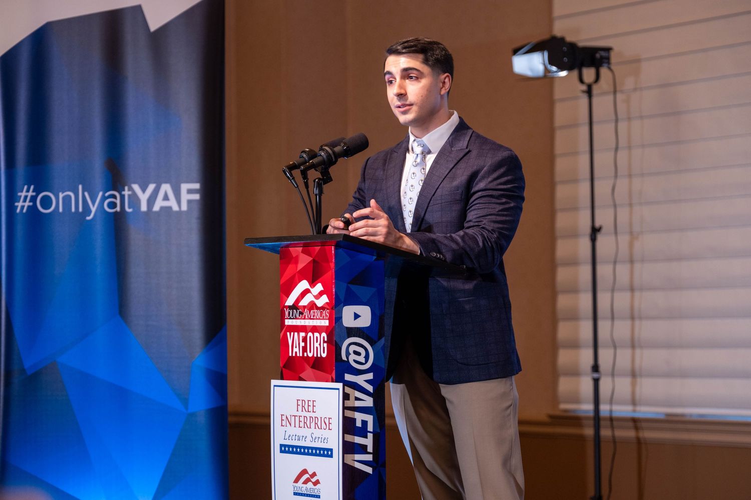 Young man in blazer speaking at YAF Free Enterprise podium at indoor event.