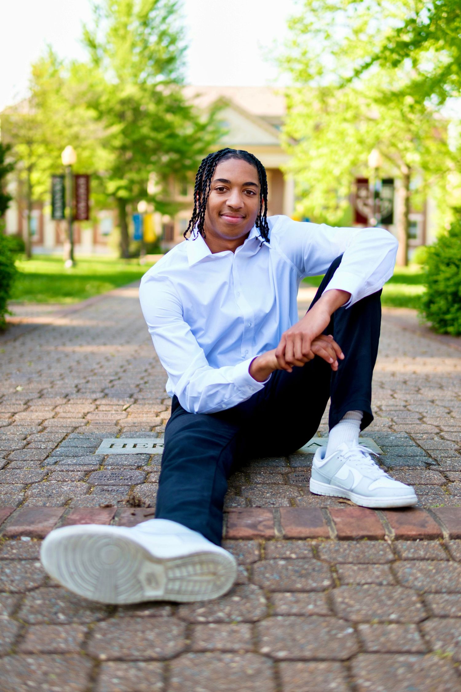 Young man with braids sitting on brick pathway in park wearing white shirt.