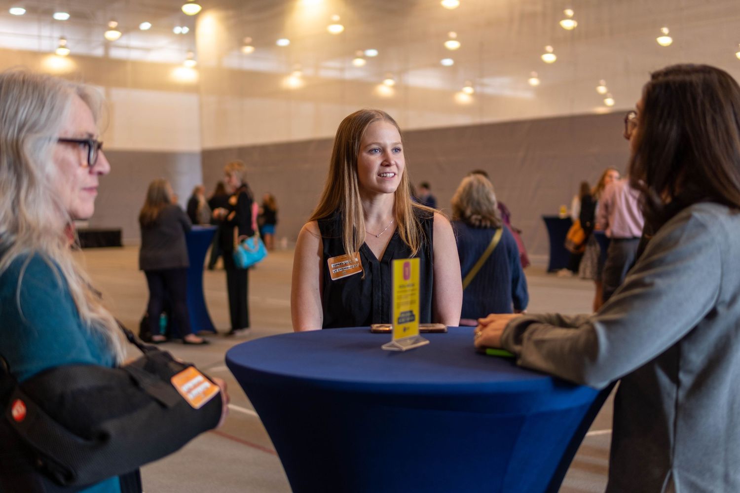 Young woman with name badge smiling and conversing with two people at a professional networking event.