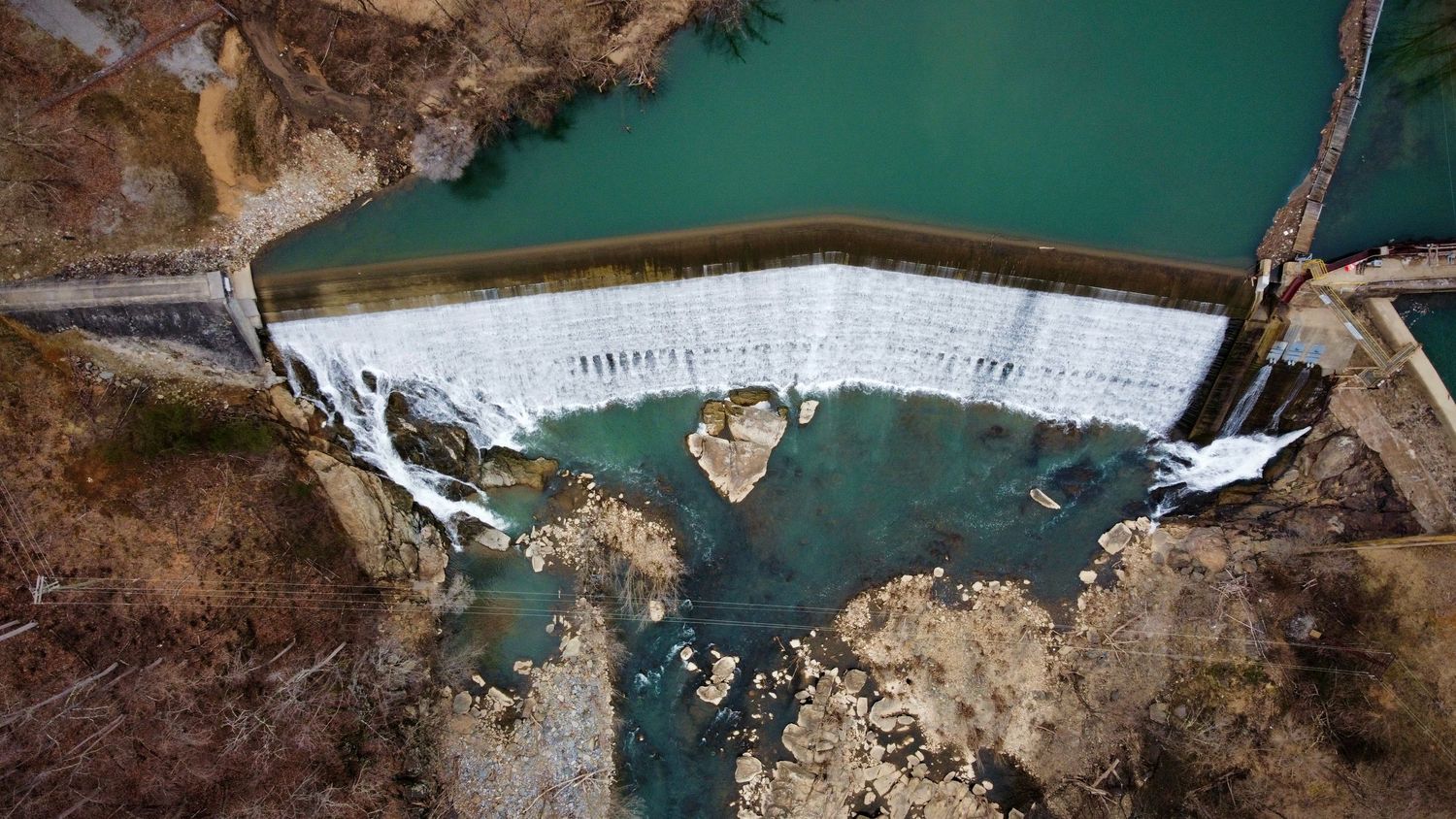 Aerial drone view of a concrete dam with turquoise water and cascading waterfall.