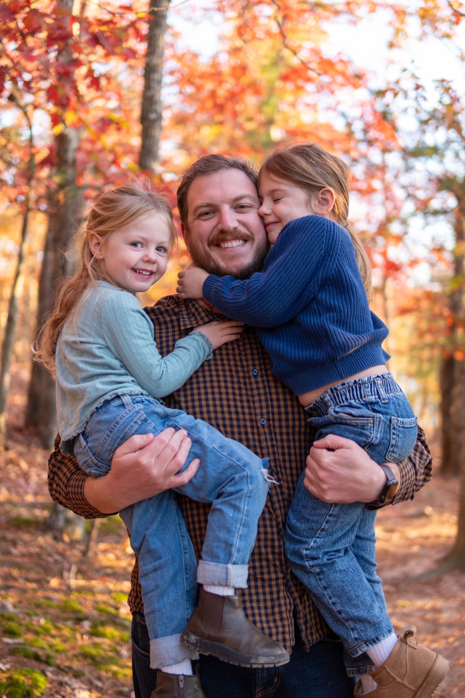 Smiling father holding two young daughters in a colorful autumn forest setting.