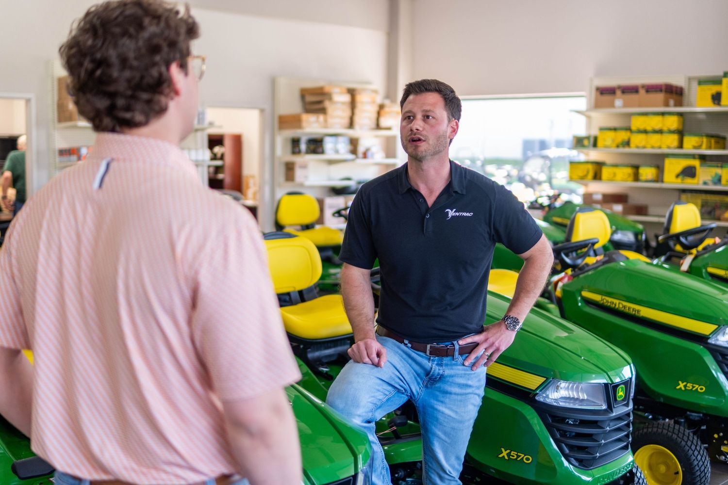 A salesman speaks with a customer in front of John Deere riding mowers inside a dealership.