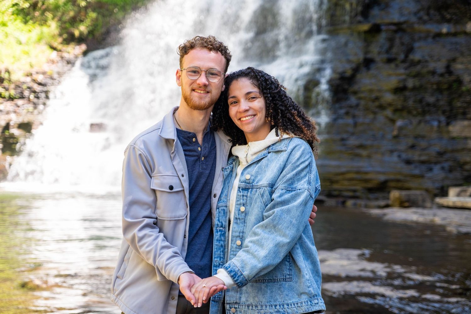 Couple holding hands smiling in front of a waterfall surrounded by lush greenery.