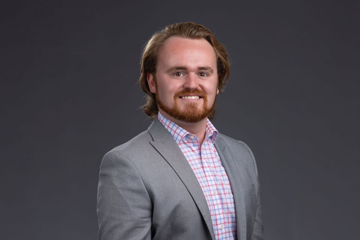 Professional headshot of young man with beard in gray blazer and plaid shirt.