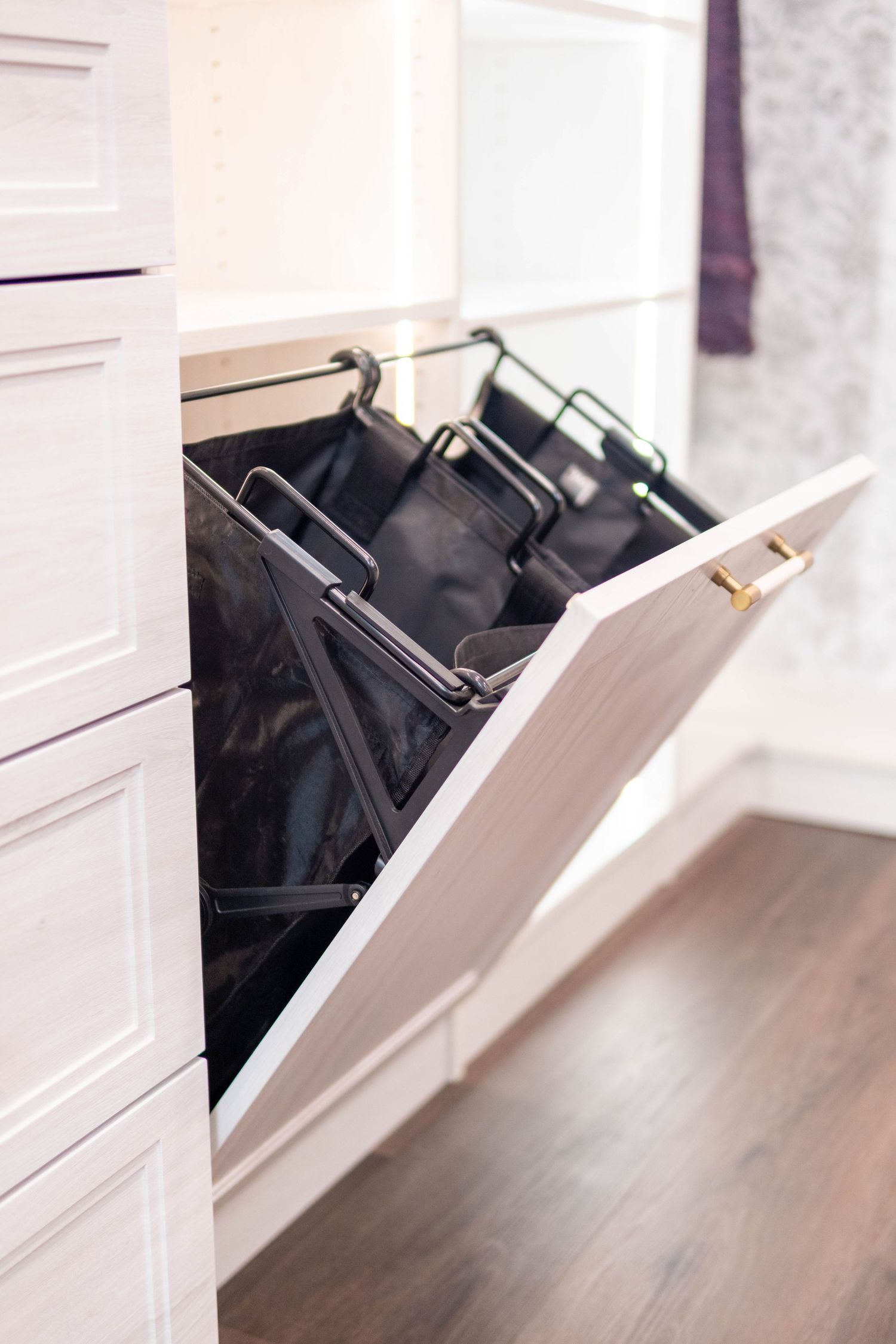 White built-in cabinet with pull-out tilt door revealing three black fabric laundry hamper bags on metal frames.