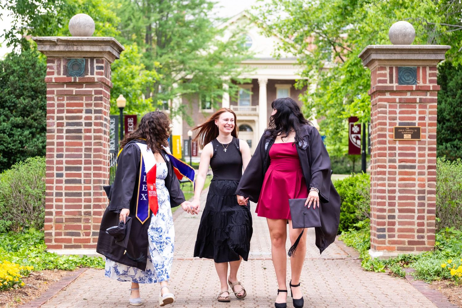 Three college graduates in caps and gowns celebrate near brick campus gates surrounded by lush green trees.