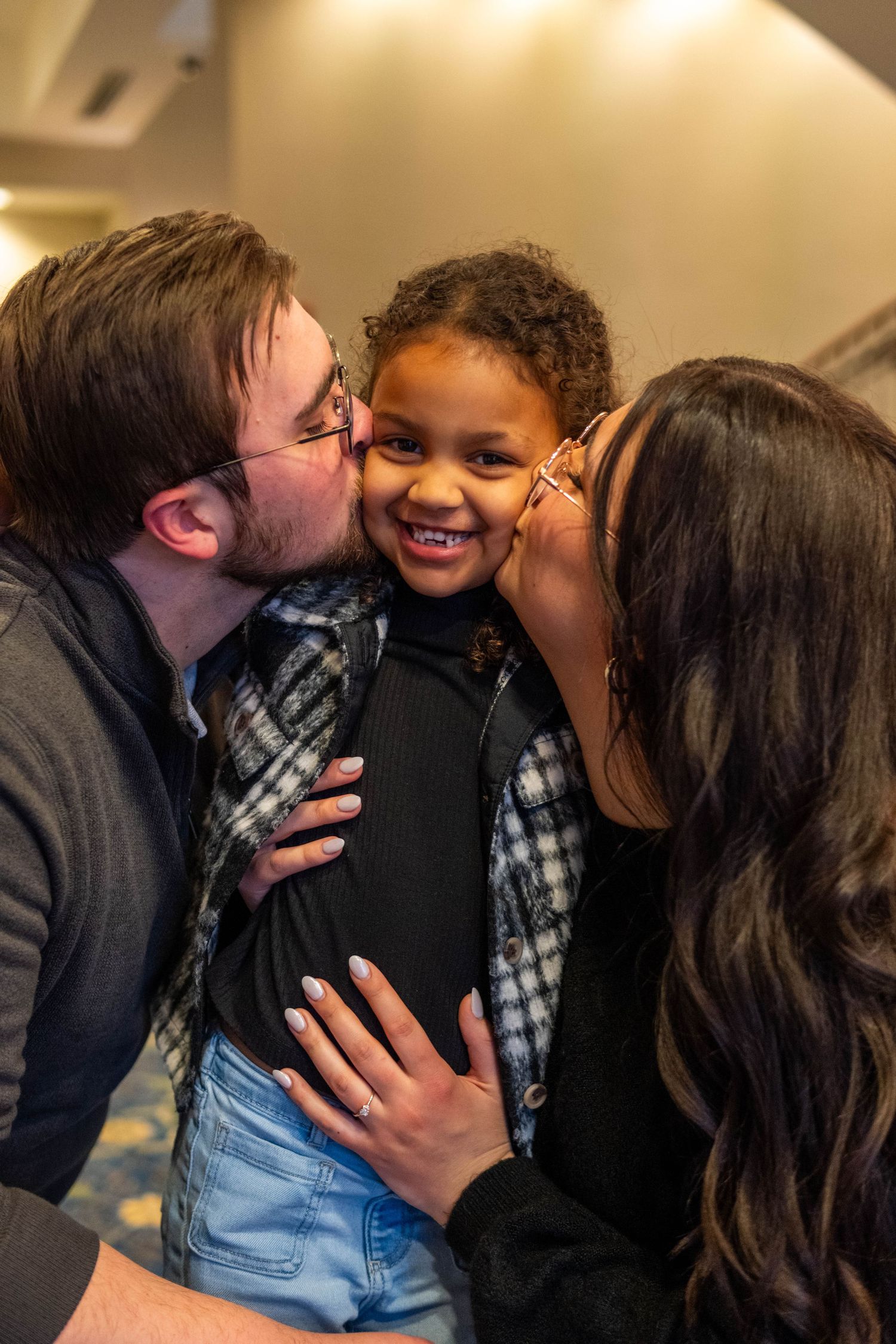 Joyful family moment with couple kissing young curly-haired child on cheeks in warmly lit interior.