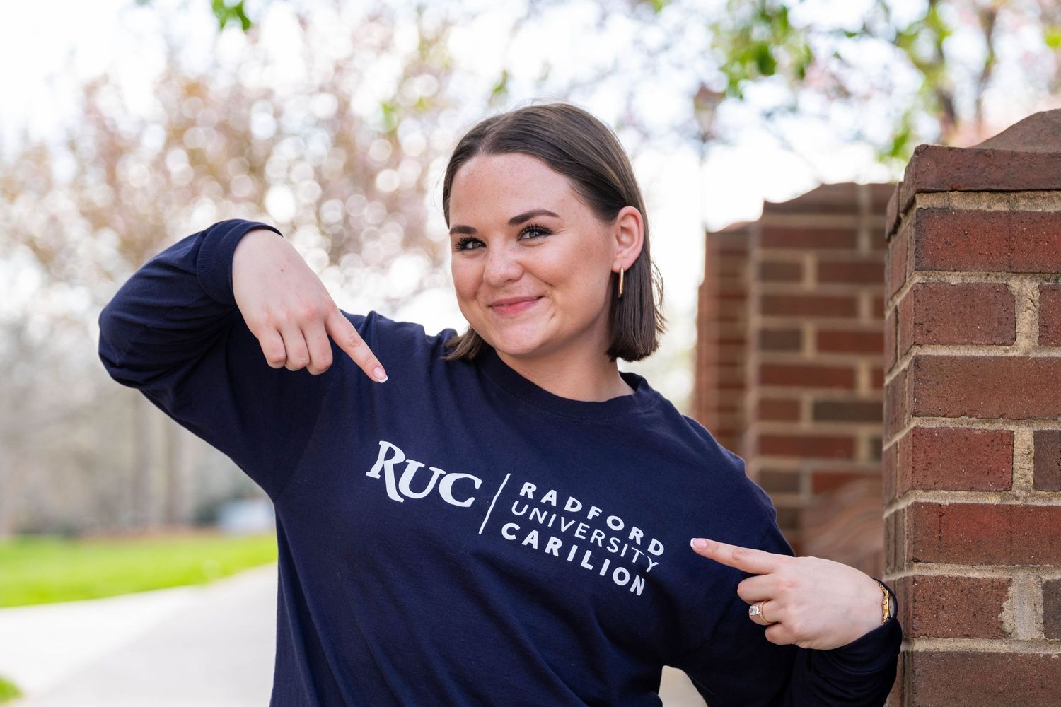 Smiling woman pointing to RUC Radford University Carilion navy blue sweatshirt outdoors.