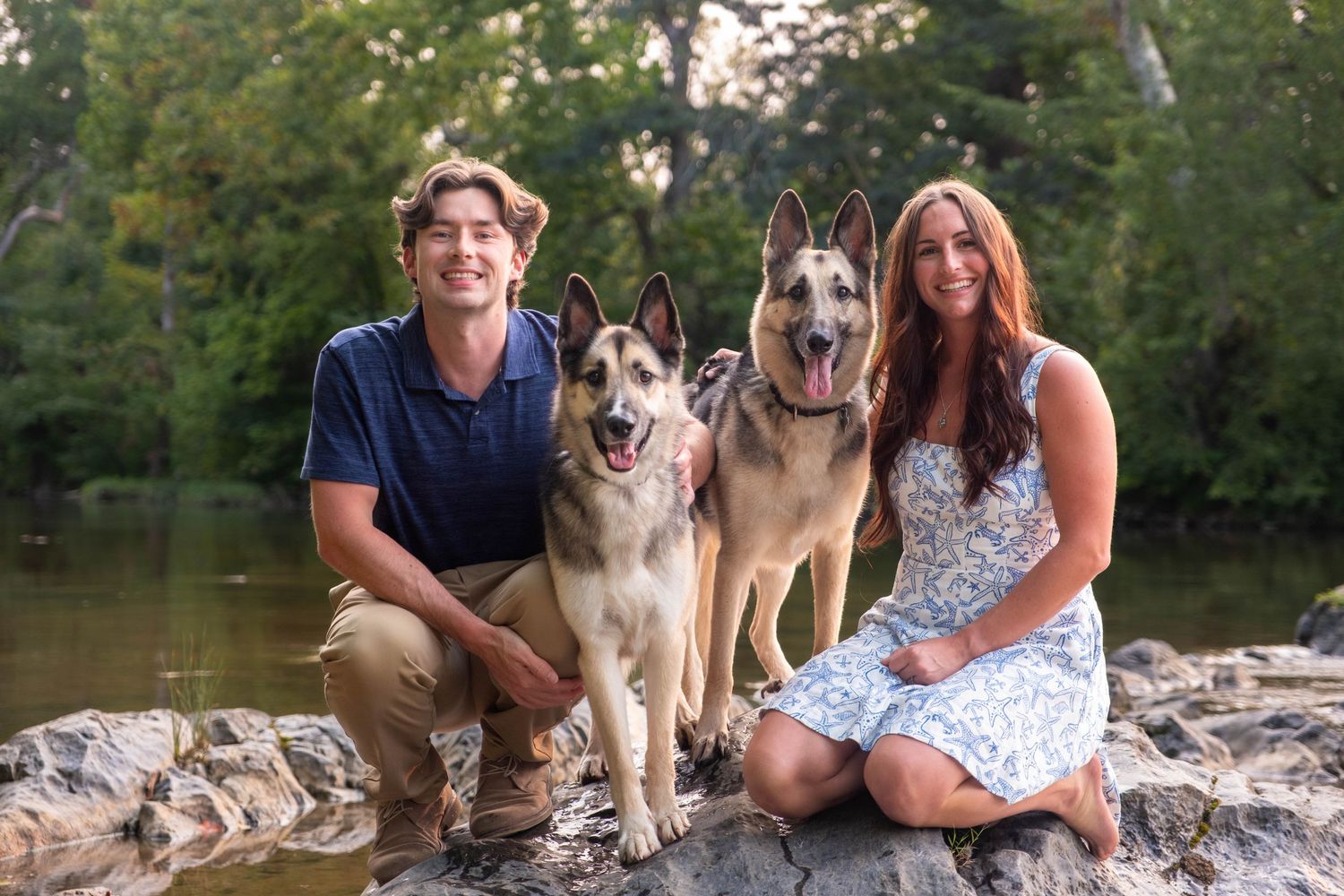 Young couple posing with two German Shepherds on rocks beside a tree-lined river.