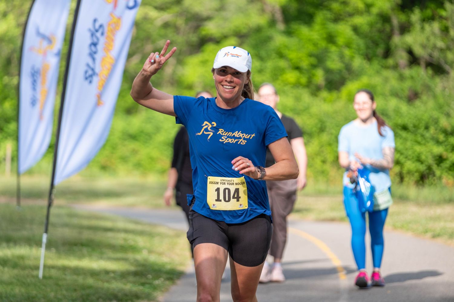 Woman wearing bib number 104 gives peace sign while running a road race on a paved path outdoors.