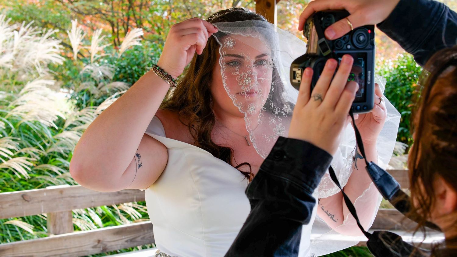 A bride adjusts her veil while a photographer captures the moment in a lush outdoor garden setting.