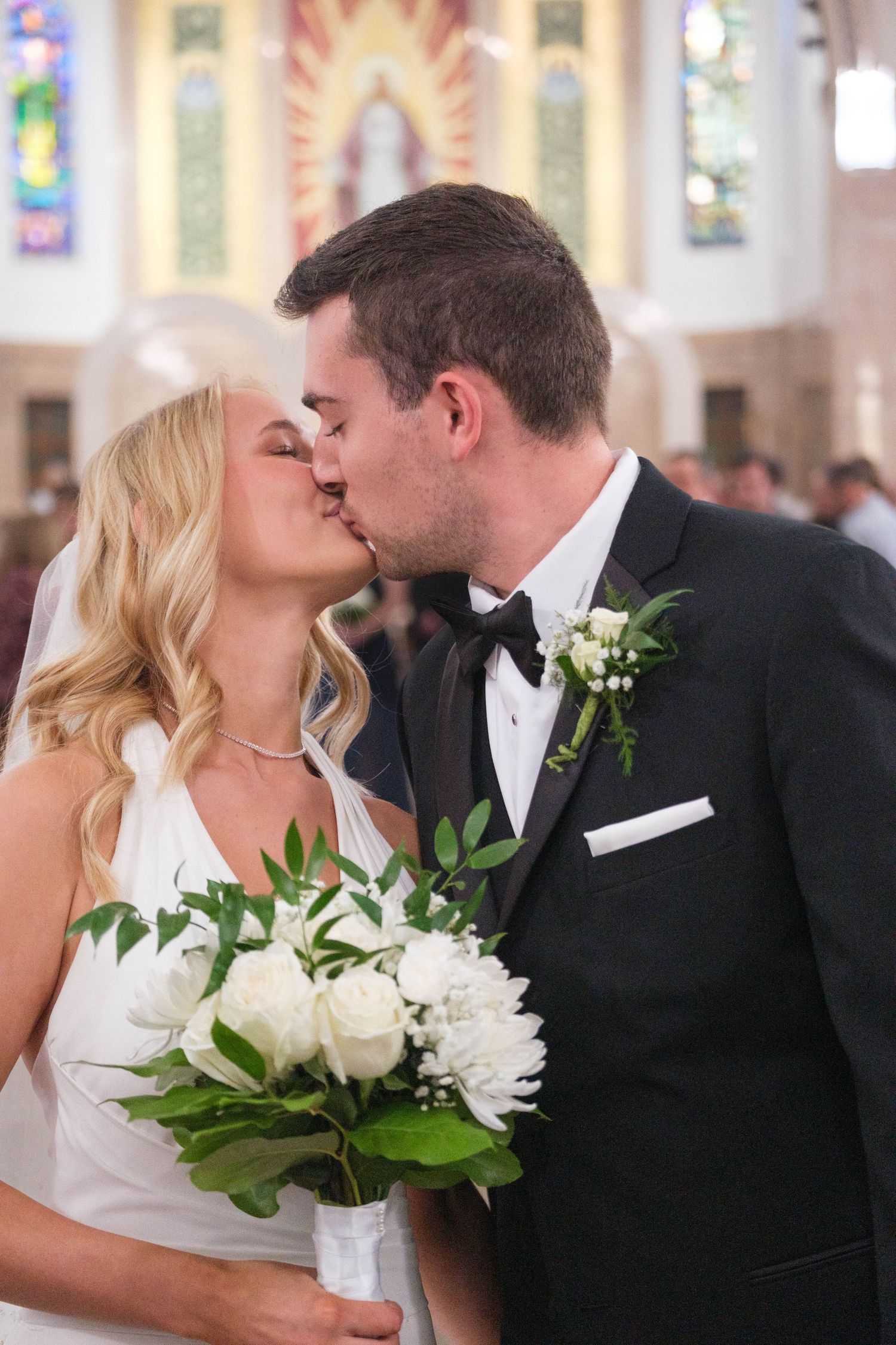 Bride and groom sharing their first kiss holding white roses bouquet inside a beautiful church.