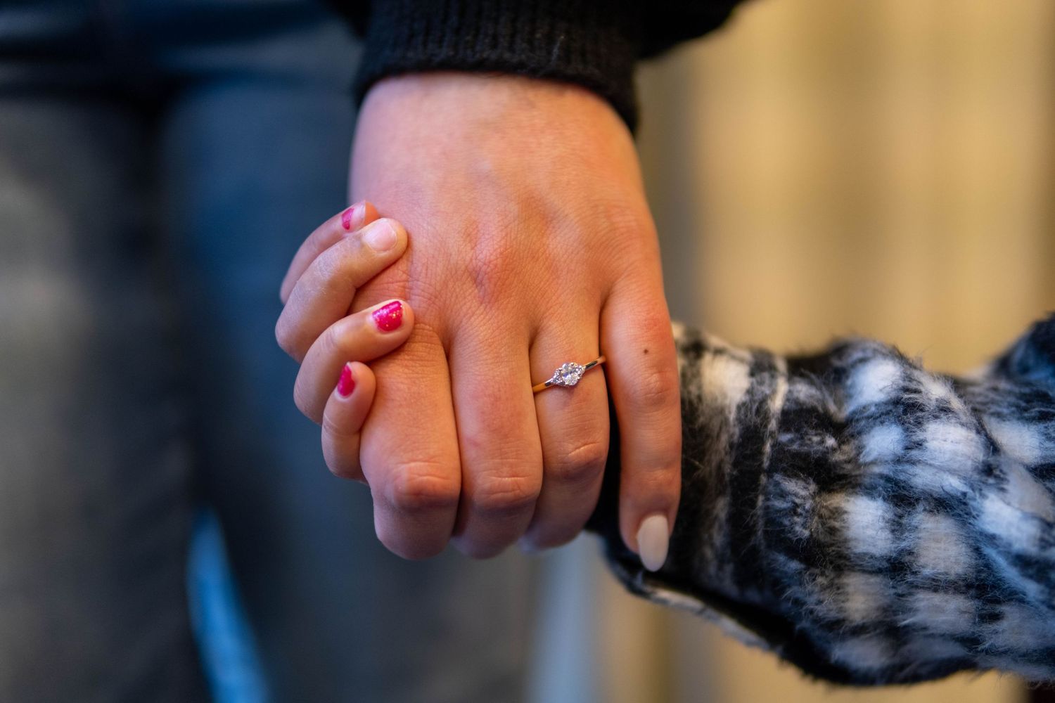 Couple holding hands showing diamond engagement ring, wearing casual clothing with plaid jacket detail.