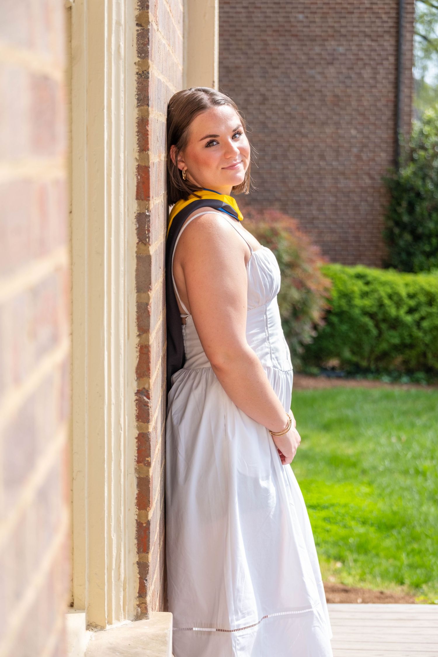Smiling woman in white dress and graduation hood leaning against column with brick wall background.