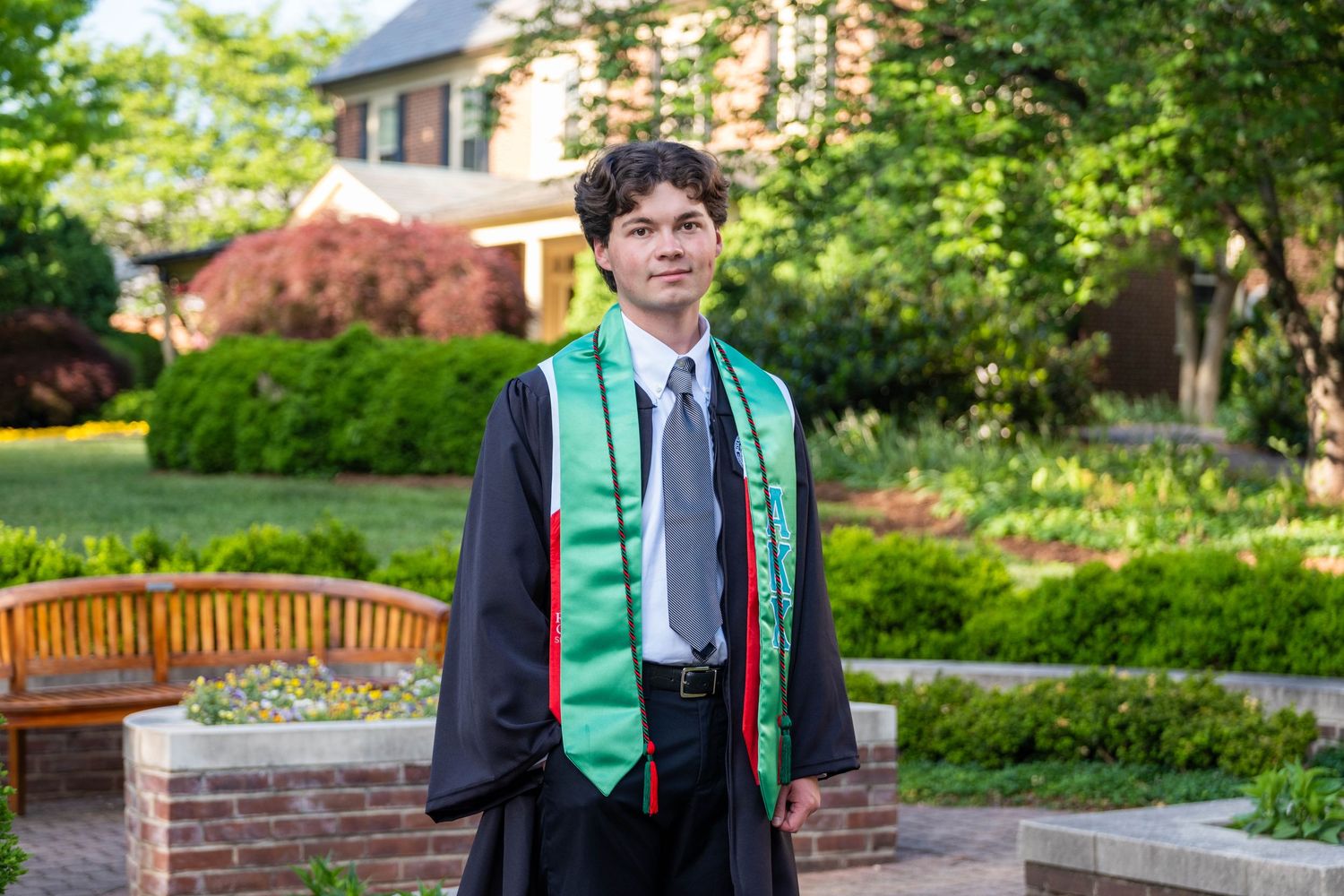 Young man in graduation gown with green stole poses outdoors on brick path with lush garden background.