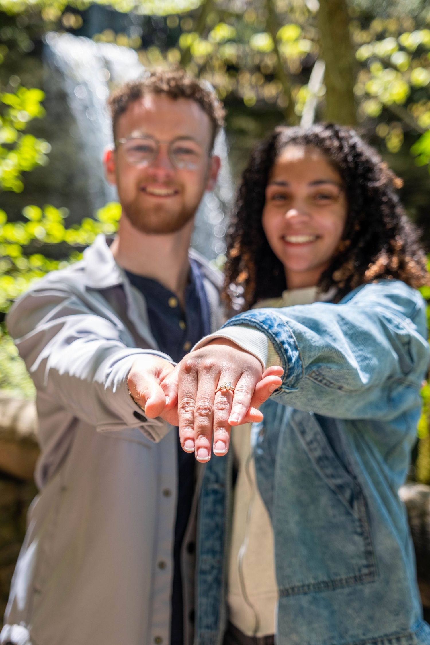 Happy couple showing off engagement ring outdoors near waterfall, smiling and extending hands toward camera.
