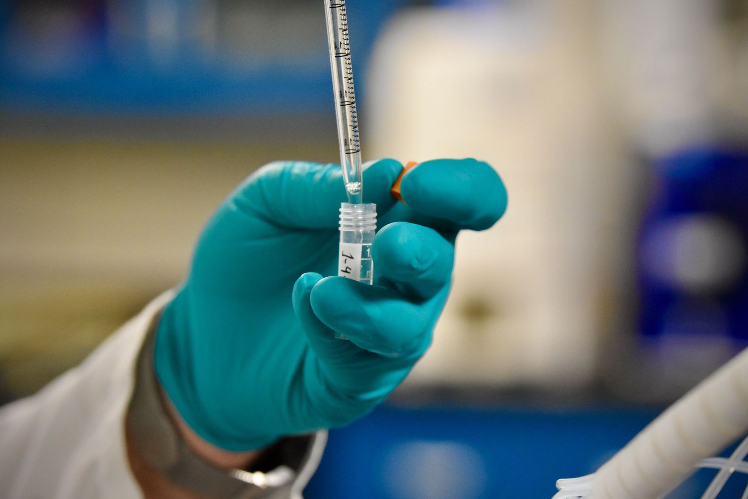 Teal gloved hand inserting a needle into a medical vial in a laboratory setting.
