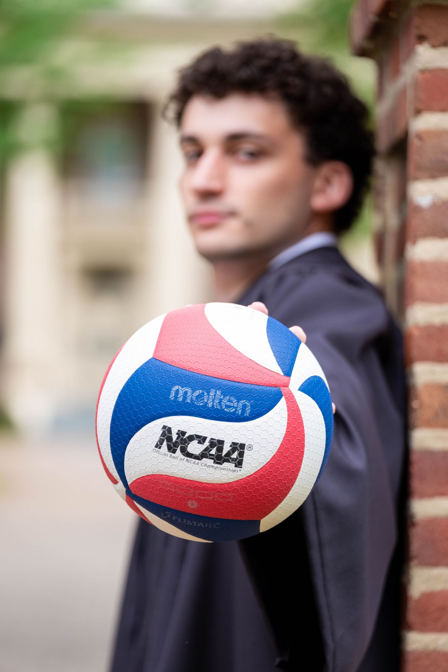 Young man in suit holding a Molten NCAA volleyball toward camera near brick wall outdoors.