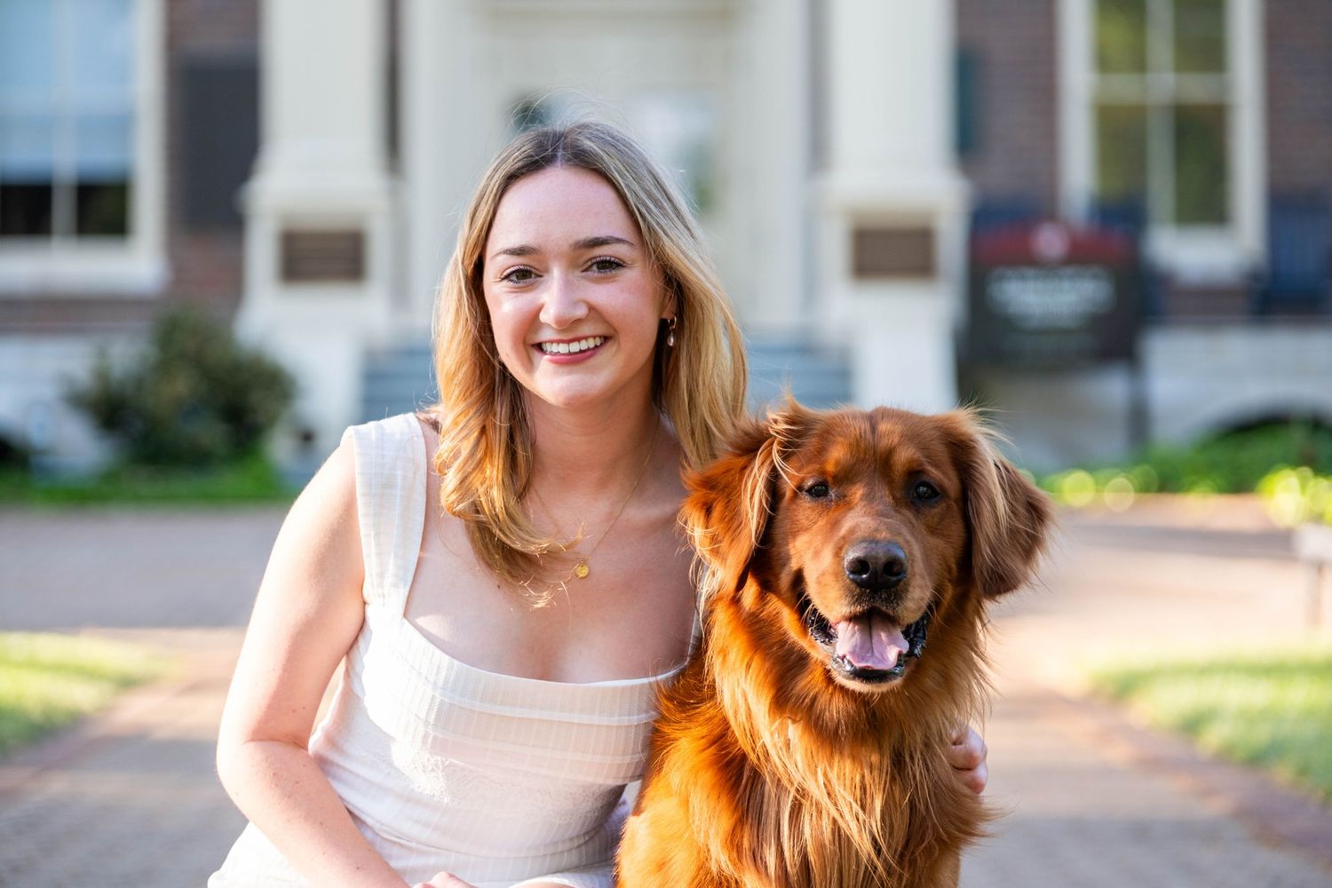 Young woman smiling with a golden retriever dog outdoors in front of a brick building on a sunny day.