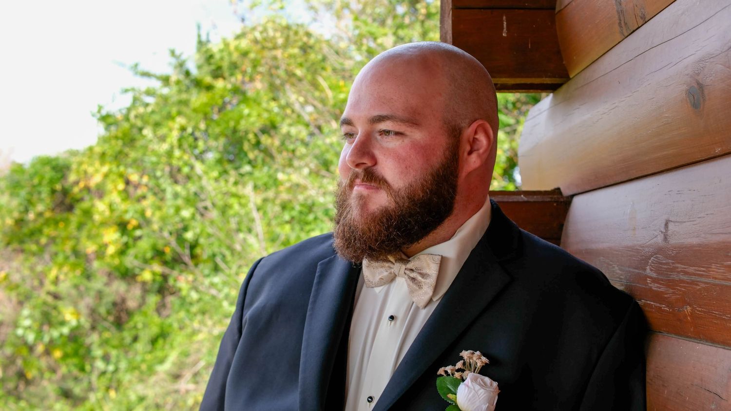 A bearded groom in a navy suit with a floral boutonniere leans against a log cabin wall outdoors.