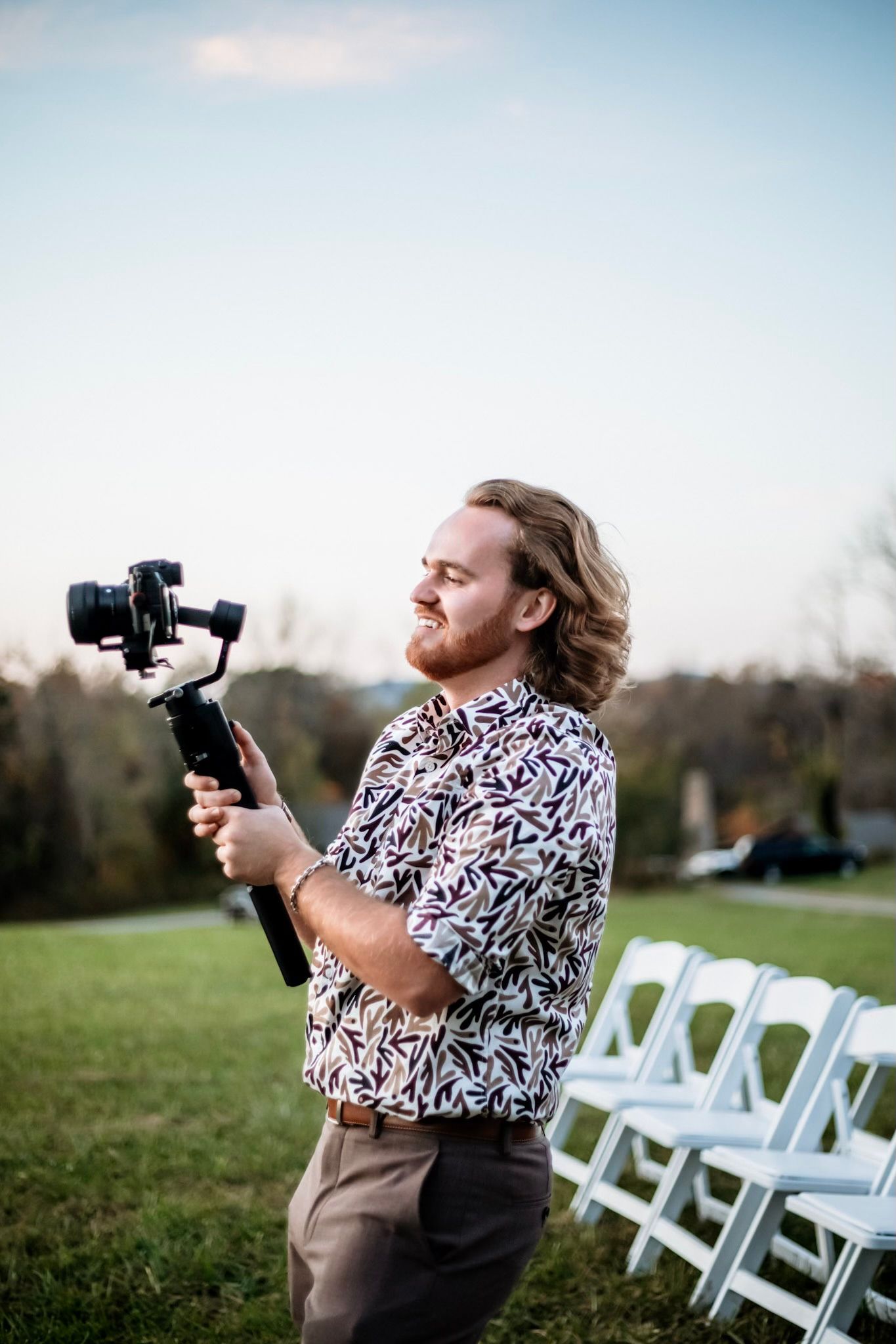 Long-haired videographer holding a gimbal stabilizer at an outdoor wedding venue with white chairs.